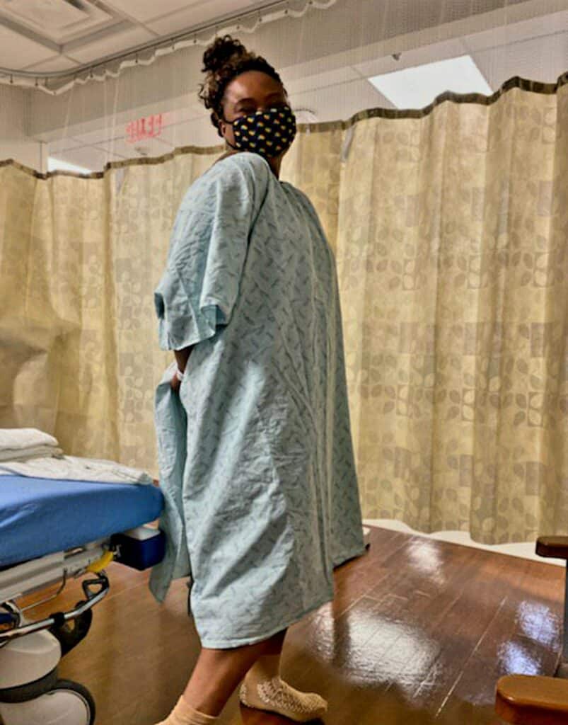  A woman in a hospital gown and mask stands with her back turned, looking over her shoulder in a curtained hospital room.