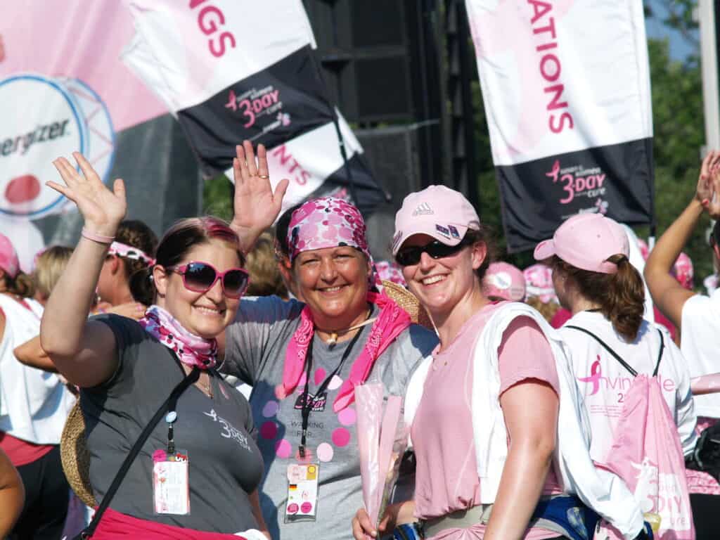Three women dressed in pink and gray, some wearing bandanas and hats, smile and wave at the camera during a breast cancer awareness event. They are surrounded by other participants and pink banners in the background.
