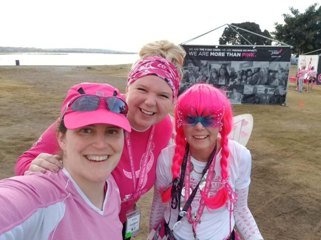 Three smiling women are dressed in pink-themed attire for a breast cancer awareness event. One woman has bright pink hair and butterfly wings. They are outdoors near a lake, with a "We Are More Than Pink" banner visible in the background.
