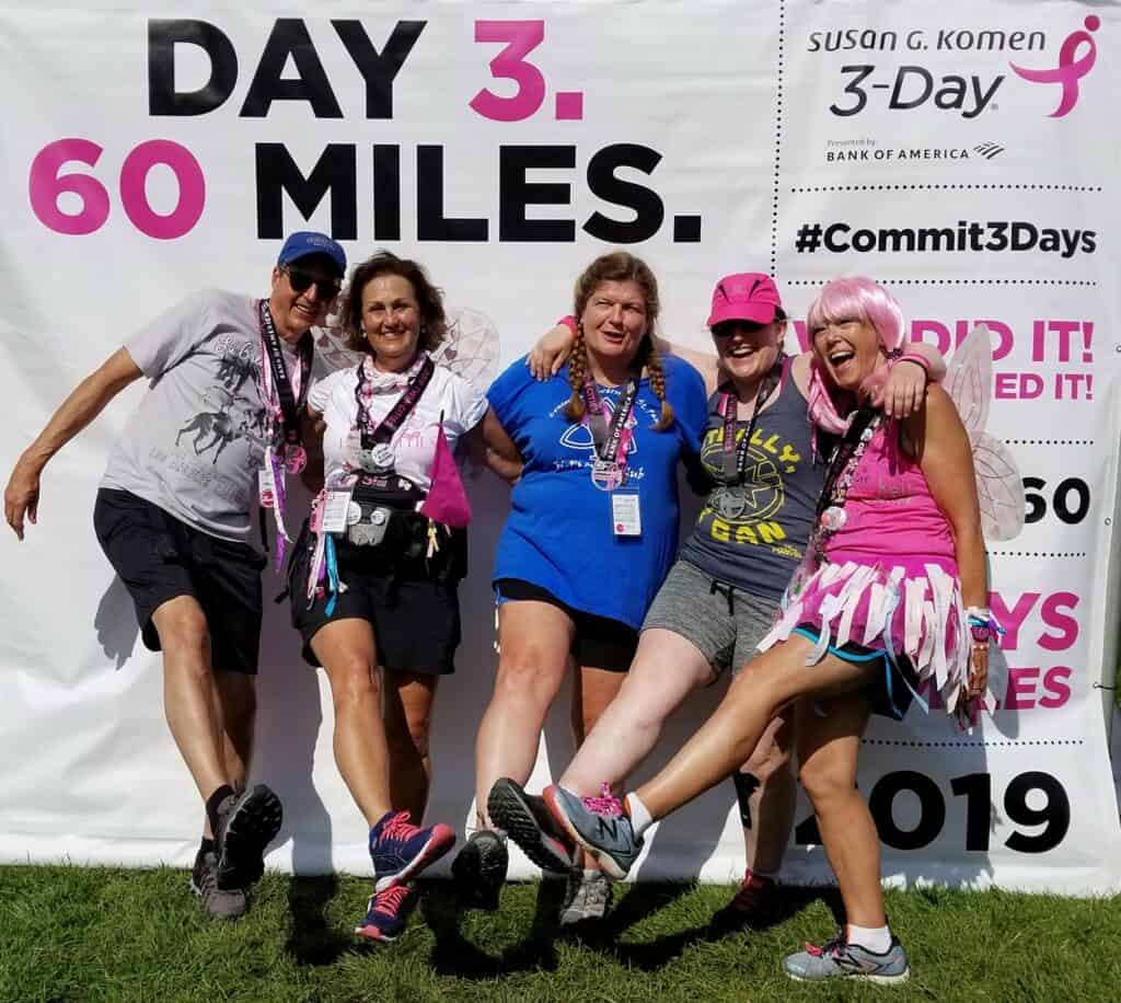 Five people smiling and posing in front of a promotional banner for the Susan G. Komen 3-Day event. They are wearing event gear and medals, indicating they completed the 60-mile walk. The background banner reads "Day 3. 60 Miles." and "Commit 3 Days" with a pink ribbon logo.
