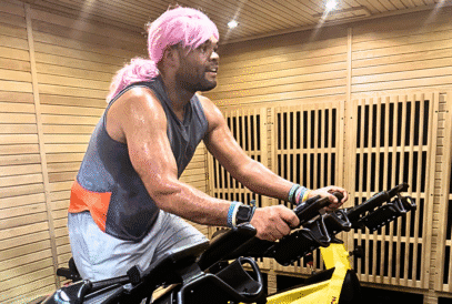 A person with a pink wig and sleeveless shirt intensely rides a stationary bike in a wooden-paneled gym, sweating and focused, as part of a breast cancer fitness fundraiser.