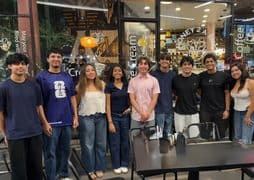 A group of nine young adults from the breast cancer awareness club stand in a line smiling in front of the glass storefront of a shop called Honey 2 A Bee.