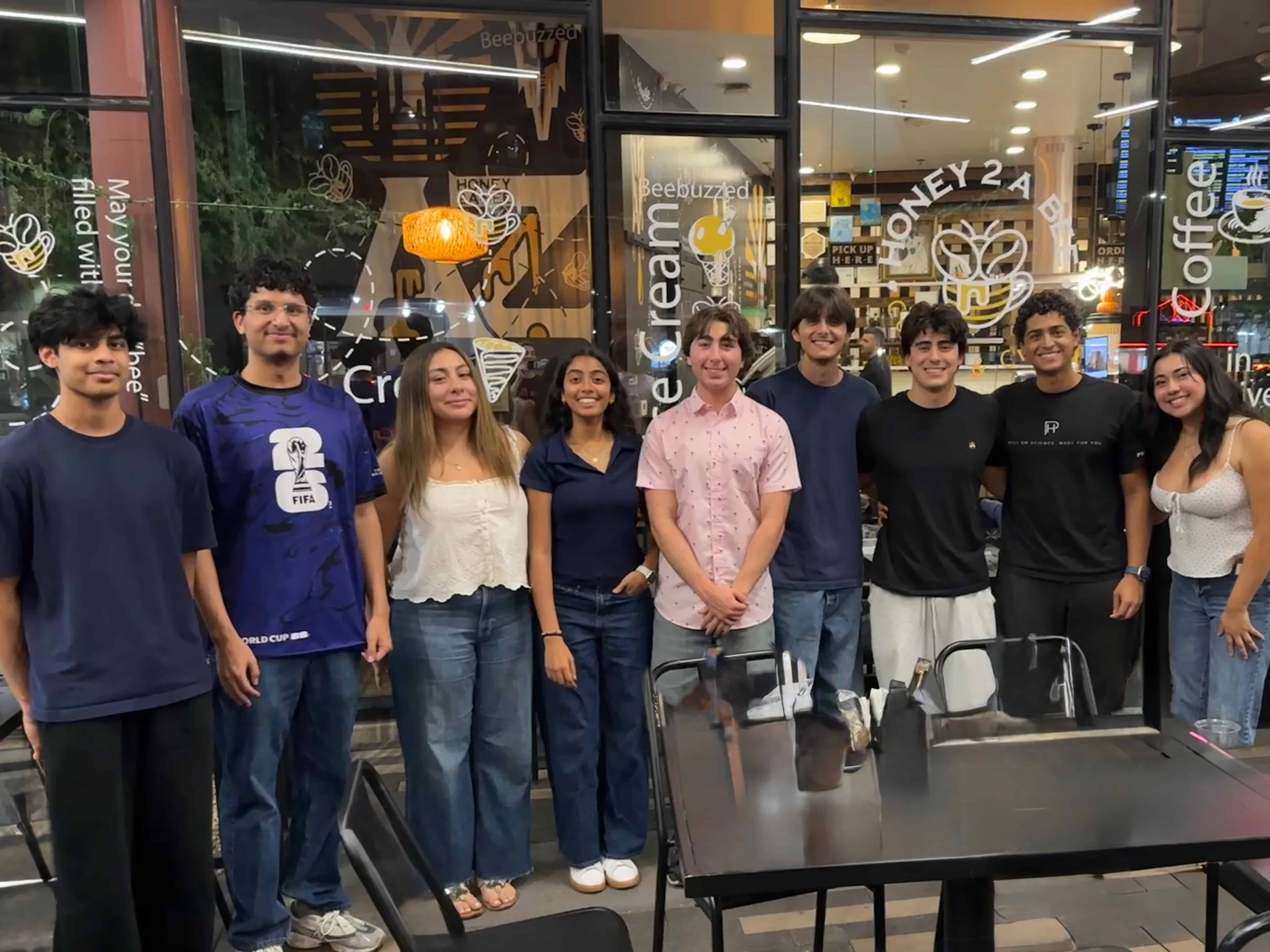 A group of nine young adults from the breast cancer awareness club stand in a line smiling in front of the glass storefront of a shop called Honey 2 A Bee.