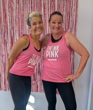 Two women wearing matching pink “Think Pink” tank tops stand side by side, smiling in front of a pink tinsel curtain at a jazzercise breast cancer fundraiser.