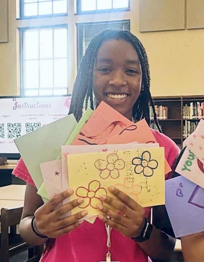 A smiling student stands in a library holding several colorful, hand-decorated cards with flowers, hearts and messages of support for breast cancer patients. Sunlight streams through large windows in the background.