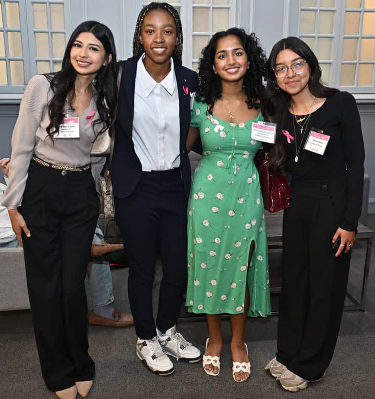 Four young women stand closely together indoors, smiling at the camera. Two wear name badges and all have pink ribbon pins, suggesting involvement in a breast cancer awareness event. They are dressed in business-casual and semi-casual attire.