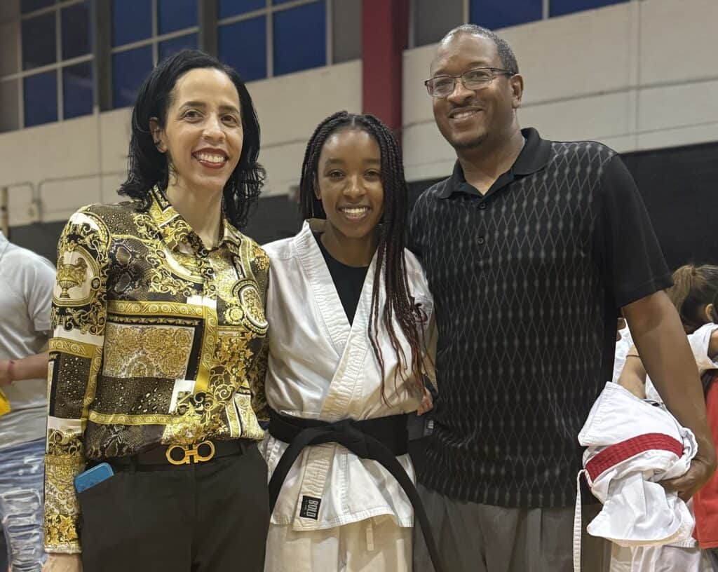 Three people smile at the camera; the woman on the left wears a patterned shirt, the young woman in the middle wears a martial arts uniform and black belt, and the man on the right wears a black patterned shirt.