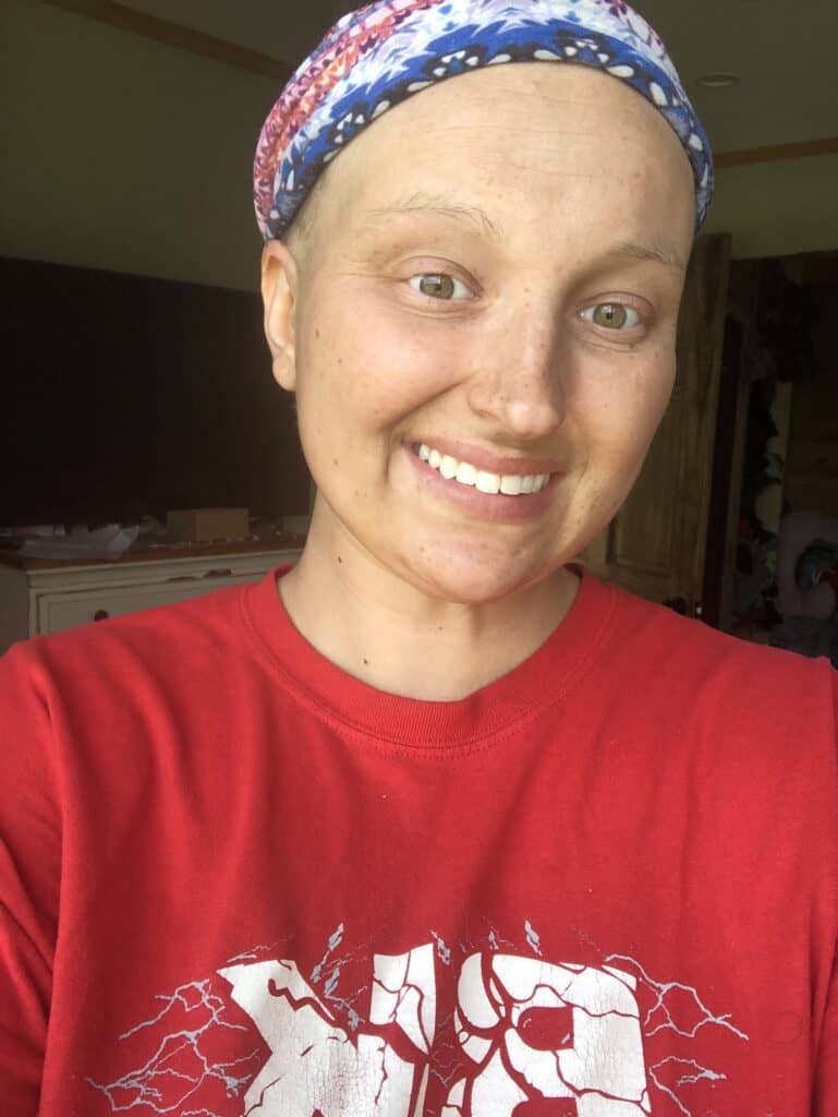 Alexis smiles at the camera. She is wearing a red shirt and her head is covered with a scarf. She has lost her hair because of breast cancer treatment.
