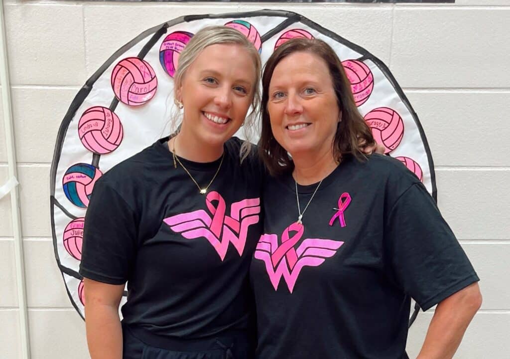 Two women wearing matching black shirts with a pink ribbon and "W" logo smile and stand together in front of a volleyball-themed display, supporting a volleyball breast cancer fundraiser with pink volleyball drawings on a white background.