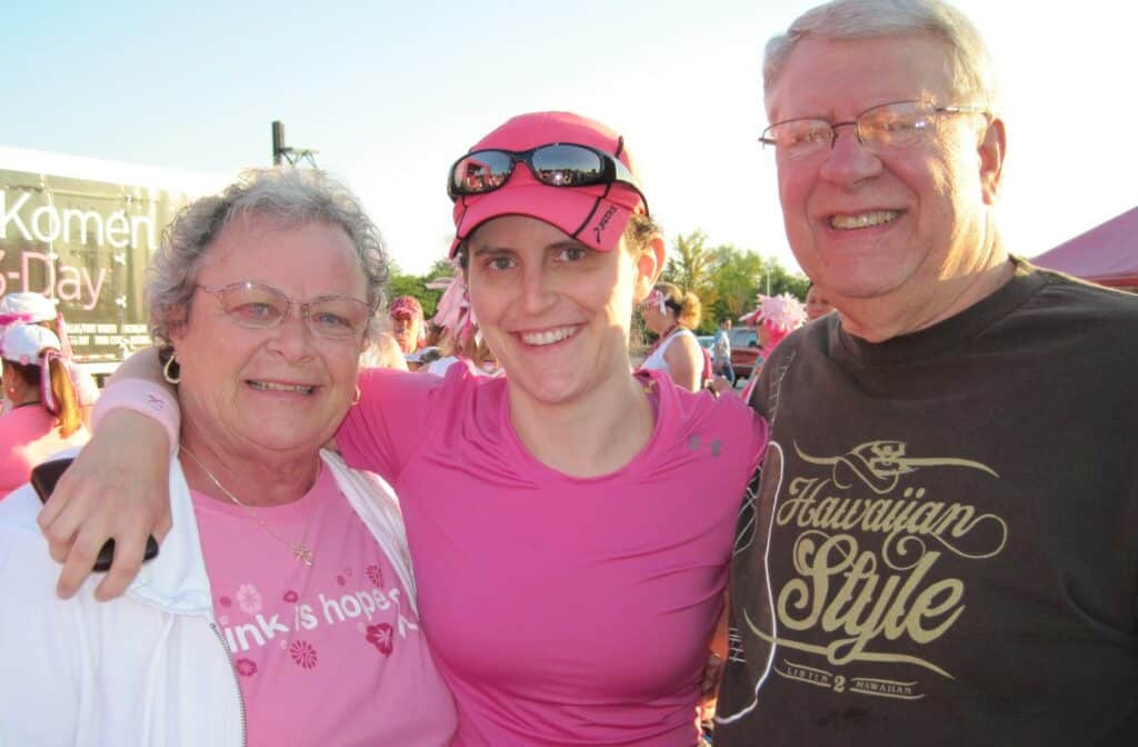 
A group of three people, two elderly individuals and a younger person in the middle, pose together, smiling at an outdoor event. They wear pink attire related to the Susan G. Komen 3-Day. A sign in the background reads "Susan G. Komen 3-Day."