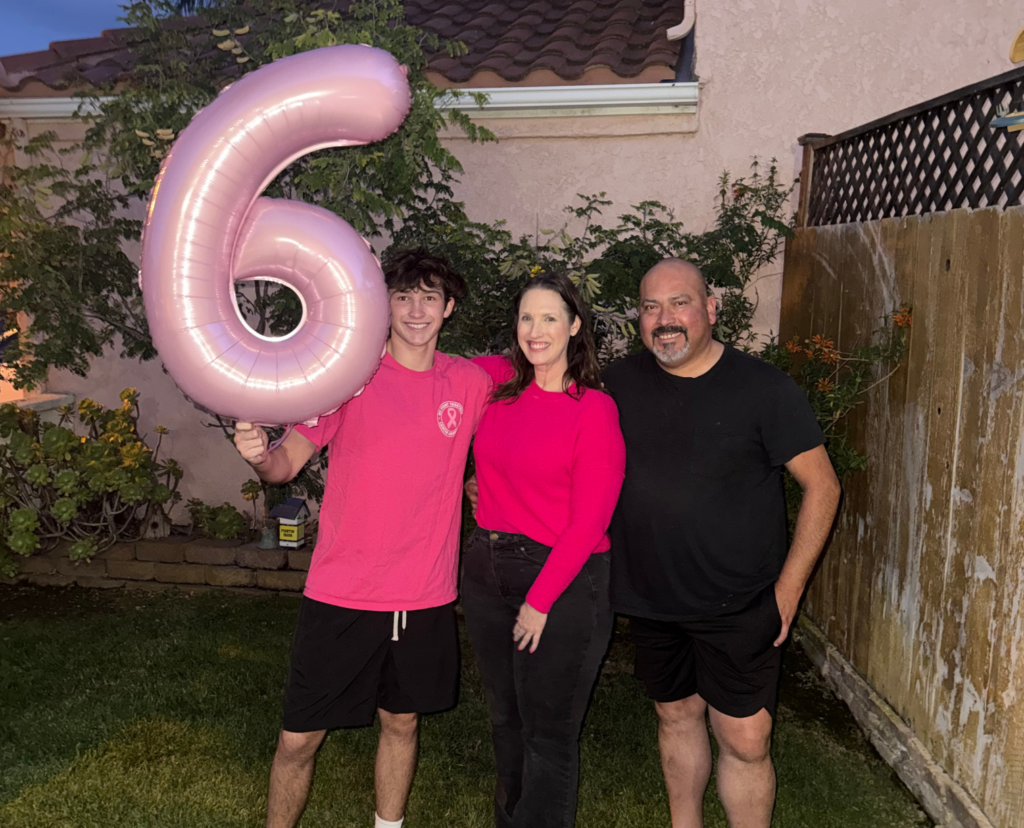 Three people stand outside, smiling. The person on the left holds a large pink balloon shaped like the number 6 – reminiscent of the pink often associated with breast cancer awareness. They stand on grass, in front of a house and wooden fence, with plants on the left.