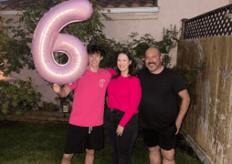 Three people stand outside, smiling. The person on the left holds a large pink balloon shaped like the number 6 – reminiscent of the pink often associated with breast cancer awareness. They stand on grass, in front of a house and wooden fence, with plants on the left.