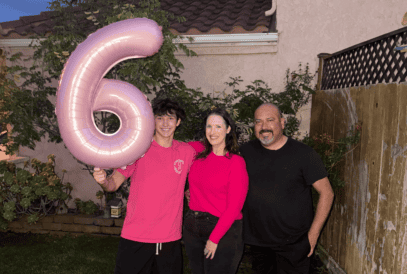 Three people stand outside, smiling. The person on the left holds a large pink balloon shaped like the number 6 – reminiscent of the pink often associated with breast cancer awareness. They stand on grass, in front of a house and wooden fence, with plants on the left.