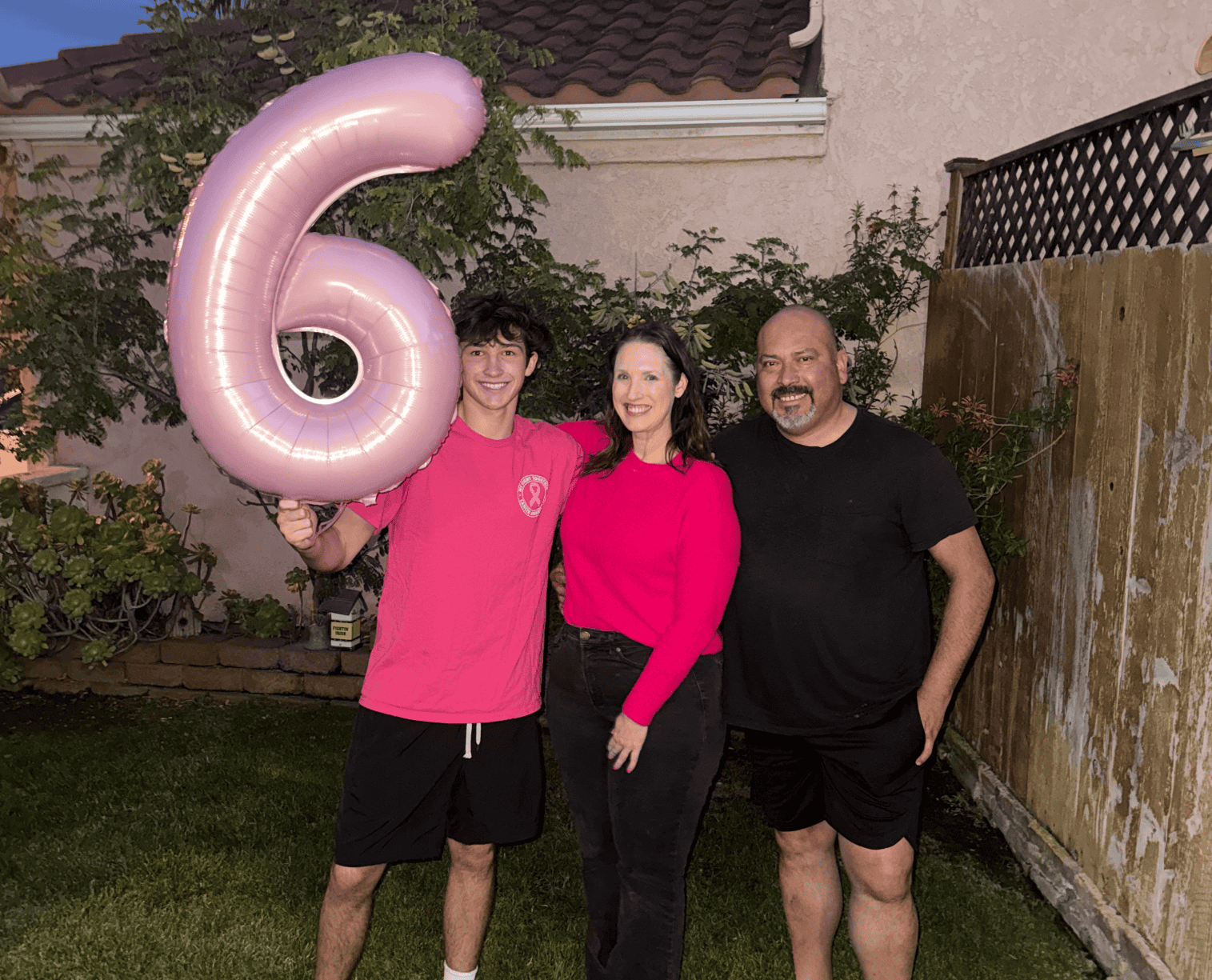 Three people stand outside, smiling. The person on the left holds a large pink balloon shaped like the number 6 – reminiscent of the pink often associated with breast cancer awareness. They stand on grass, in front of a house and wooden fence, with plants on the left.