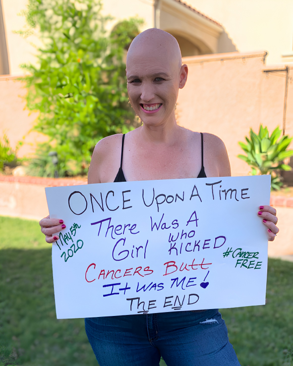 A smiling bald woman in a black tank top holds a colorful sign that says she beat breast cancer and is now cancer-free, standing outside in a sunny garden.