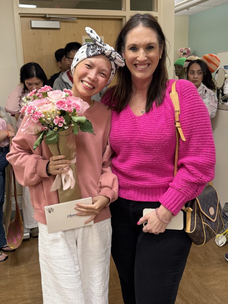 Two women smiling together indoors—one, possibly a mentor, holds pink flowers and an envelope; both wear pink tops. This touching moment likely takes place within a breast cancer support group. Other people stand in the background, some looking toward the camera.