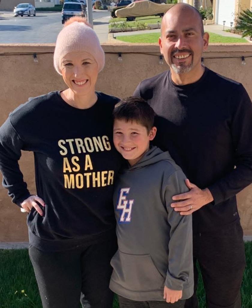 Three people pose outside on grass: a smiling woman in a pink beanie and “STRONG AS A MOTHER” sweater—showing strength and resilience in the face of breast cancer—a boy in a gray hoodie, and a smiling man in a black shirt. Bright sunlight and a suburban background.
