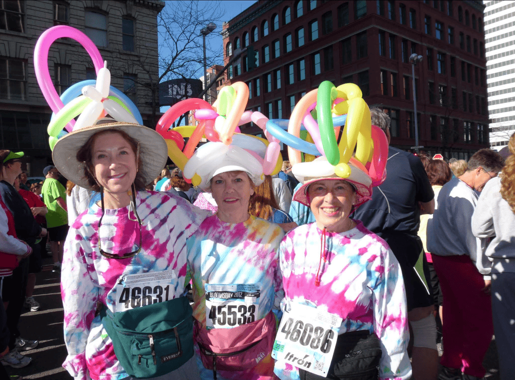 Three smiling women wear tie-dye shirts, hats, and colorful balloon headpieces at the Bloomsday breast cancer fundraiser. Each has a race bib, with a crowd and buildings visible in the background.