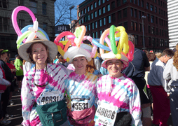 Three smiling women wear tie-dye shirts, hats, and colorful balloon headpieces at the Bloomsday breast cancer fundraiser. Each has a race bib, with a crowd and buildings visible in the background.