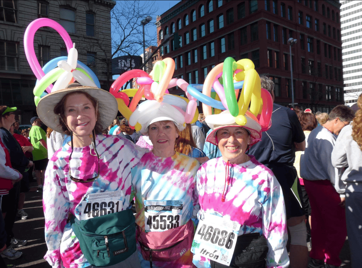 Three smiling women wear tie-dye shirts, hats, and colorful balloon headpieces at the Bloomsday breast cancer fundraiser. Each has a race bib, with a crowd and buildings visible in the background.