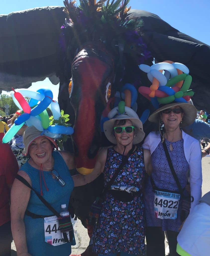 Three women in sunhats and balloon hats smile at a Bloomsday breast cancer fundraiser parade, standing before a colorful bird float. Wearing race bibs and sunglasses, they enjoy the festive, sunny atmosphere.