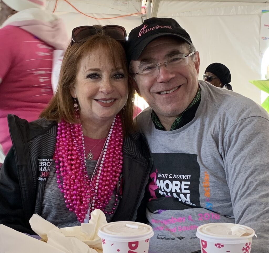 A smiling woman and man sit together at an event. Both wear "Susan G. Komen MORE THAN PINK" shirts; the woman has bright pink bead necklaces that signify the years since her breast cancer diagnosis. Pink cups and food are on the table in front of them.