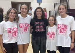 Five women and girls stand smiling in a gym, taking part in a volleyball breast cancer fundraiser. Four wear white “Pink Out” shirts supporting awareness, while the woman in the center wears a black shirt with pink text.