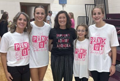 Five women and girls stand smiling in a gym, taking part in a volleyball breast cancer fundraiser. Four wear white “Pink Out” shirts supporting awareness, while the woman in the center wears a black shirt with pink text.