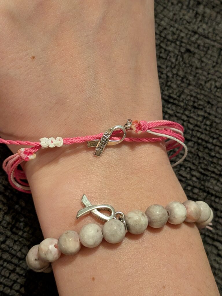 A hand with two bracelets sold as a part of a youth breast cancer fundraiser: one is pink string with a silver "hope" ribbon charm and small white beads, the other is a beaded bracelet with grayish stones and a silver ribbon charm, shown against a dark textured background.