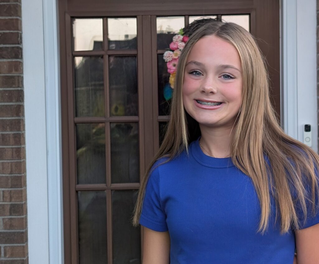 A teenage girl with long straight blonde hair and braces, wearing a blue t-shirt, stands and smiles in front of a brown door with glass panels and a floral wreath.