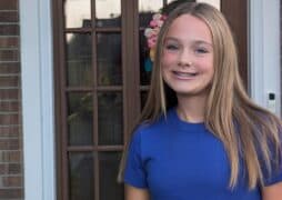 A teenage girl with long straight blonde hair and braces, wearing a blue t-shirt, stands and smiles in front of a brown door with glass panels and a floral wreath.