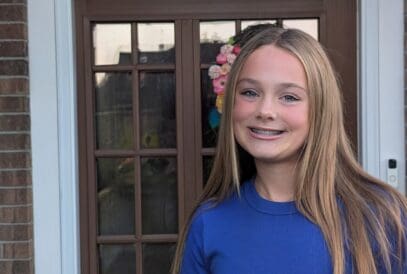 A teenage girl with long straight blonde hair and braces, wearing a blue t-shirt, stands and smiles in front of a brown door with glass panels and a floral wreath.