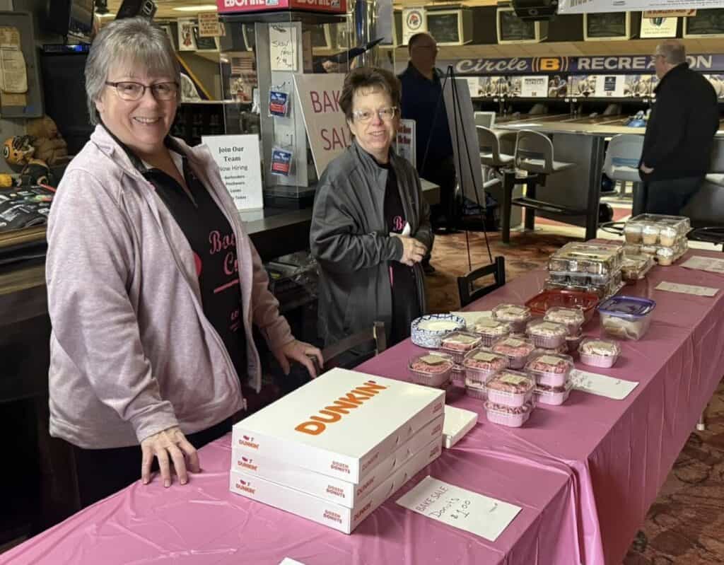 Two women stand behind a table covered with a pink cloth, displaying baked goods and Dunkin’ donut boxes at a bake sale inside a recreational venue. Signs and treats are arranged neatly on the table.