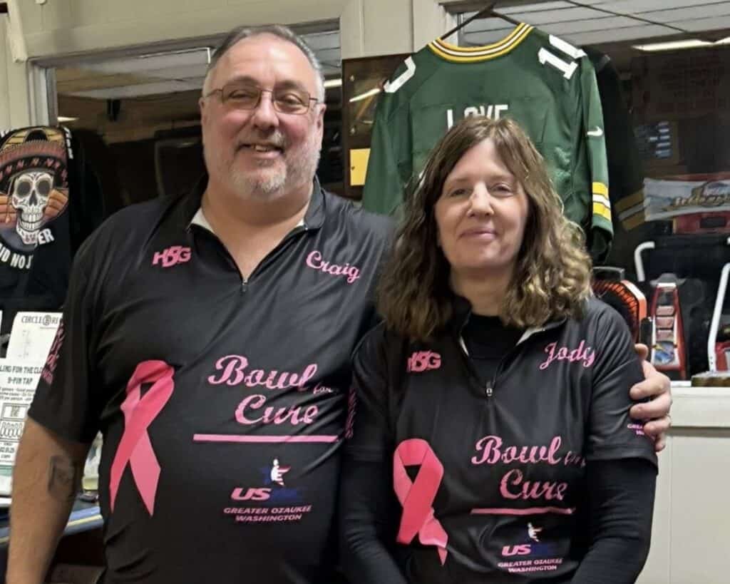 A man and a woman stand side by side, smiling, wearing matching “Bowl for the Cure” shirts with pink ribbons, showing their support for the breast cancer bowling fundraiser. Bowling memorabilia and a green jersey are visible in the background.