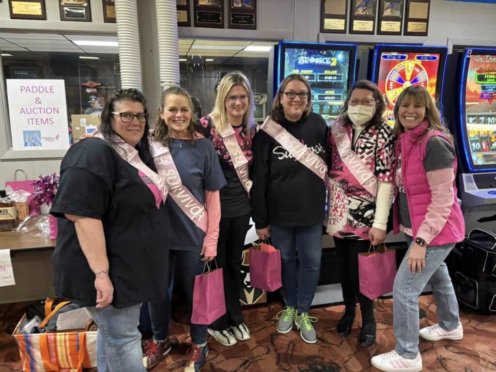 Six women stand together indoors, some wearing pink "Survivor" sashes and holding pink gift bags. They are smiling, with arcade games and a sign for paddle and auction items in the background.