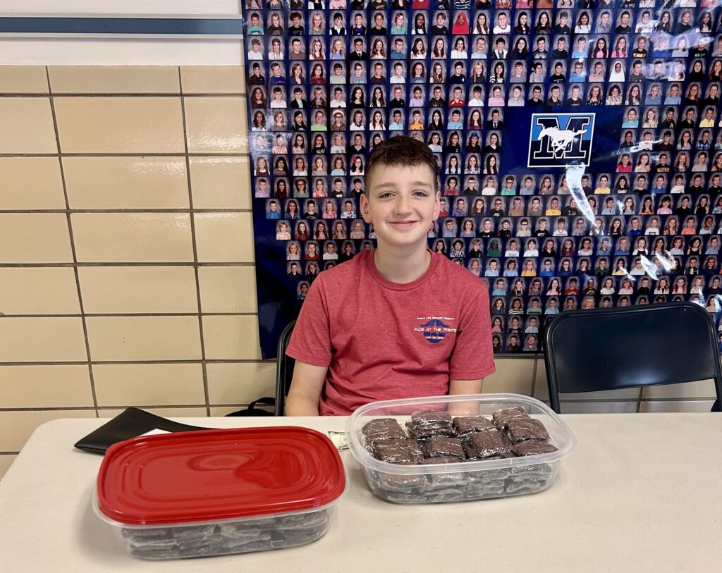 A boy in a red shirt sits at a table with containers of brownies in front of him, smiling at a breast cancer bake sale fundraiser. Behind him is a wall with many small portraits and a school logo.