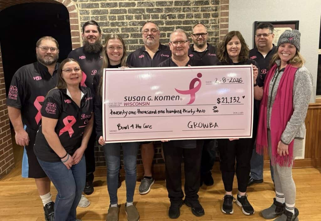 A group of eleven people holds a large pink check for $21,132 made out to Susan G. Komen Wisconsin from the "Bowl for the Cure" breast cancer bowling fundraiser. Most wear matching black shirts with pink ribbons, celebrating their impact.