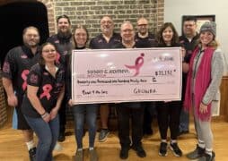 A group of eleven people holds a large pink check for $21,132 made out to Susan G. Komen Wisconsin from the "Bowl for the Cure" breast cancer bowling fundraiser. Most wear matching black shirts with pink ribbons, celebrating their impact.