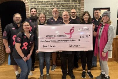A group of eleven people holds a large pink check for $21,132 made out to Susan G. Komen Wisconsin from the "Bowl for the Cure" breast cancer bowling fundraiser. Most wear matching black shirts with pink ribbons, celebrating their impact.