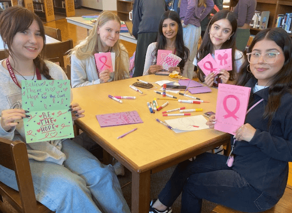 Five young women sit around a table in a classroom, holding handmade cards with pink ribbons and hopeful messages for breast cancer awareness. Art supplies are scattered on the table.