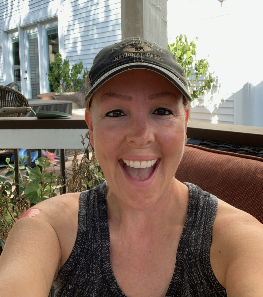 A woman wearing a black tank top and a National Park cap, smiling widely, takes a selfie outdoors on a porch filled with plants and patio furniture, proudly displaying her strength as a breast cancer survivor.