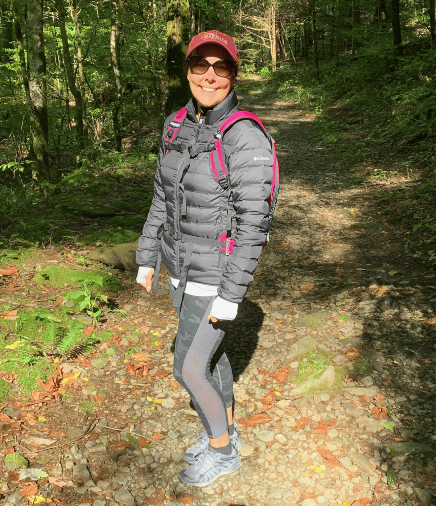 A woman in sunglasses, a maroon cap, and a black puffer jacket stands smiling on a rocky forest trail. She carries a pink-accented backpack and wears leggings and sneakers, surrounded by trees and sunlight.