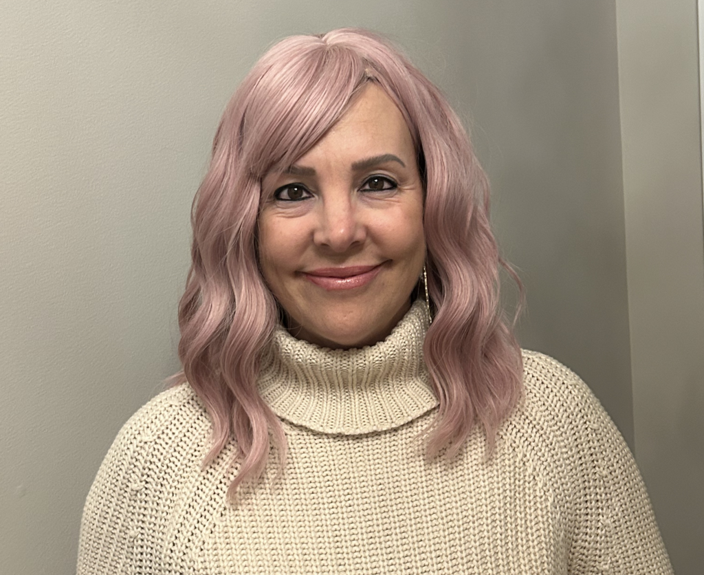 A woman wearing a wavy, pastel pink wig is smiling. As a VIPink Ambassador fundraising supporter, she wears a cream-colored, chunky knit turtleneck sweater and stands against a plain, light-colored background.
