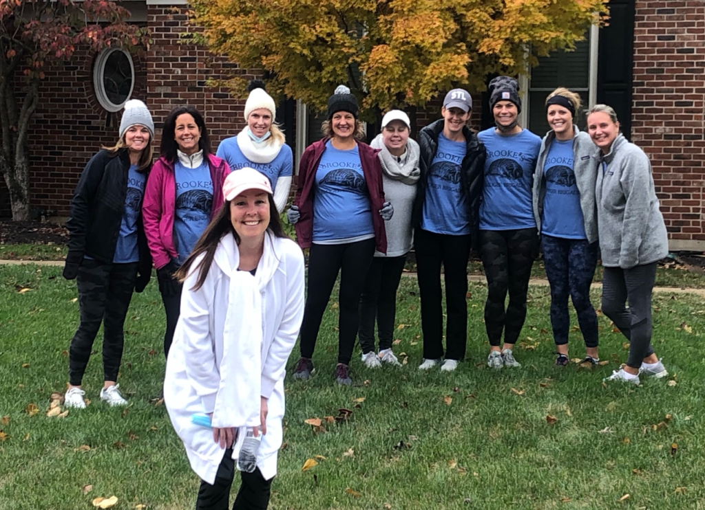 A group of nine women stands on grass in front of a tree with autumn leaves and a brick building, all wearing blue matching shirts and beanies. Another woman in white stands smiling in the foreground.
