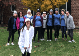 A group of nine women stands on grass in front of a tree with autumn leaves and a brick building, all wearing blue matching shirts and beanies. Another woman in white stands smiling in the foreground.