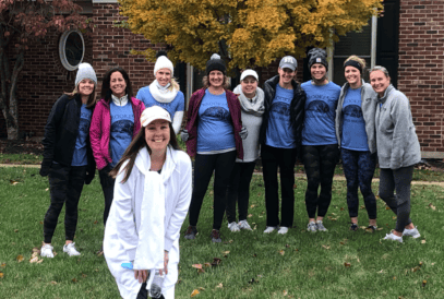 A group of nine women stands on grass in front of a tree with autumn leaves and a brick building, all wearing blue matching shirts and beanies. Another woman in white stands smiling in the foreground.