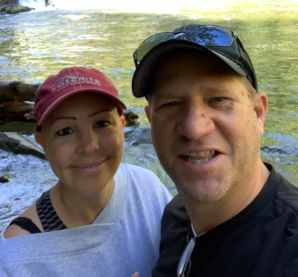 A man and woman smile for a selfie by a river with sunlight reflecting on the water. The woman wears a red cap and gray top; the man wears a black t-shirt, black cap, and sunglasses on his head. Trees are visible in the background.