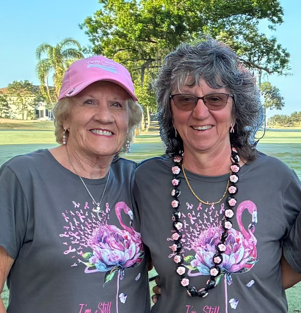 Two smiling women stand together outdoors at a golf breast cancer fundraiser, wearing matching gray shirts with pink floral designs and “I’m Still Standing – Breast Cancer Awareness” printed on them. One wears a pink hat; both look happy and supportive.