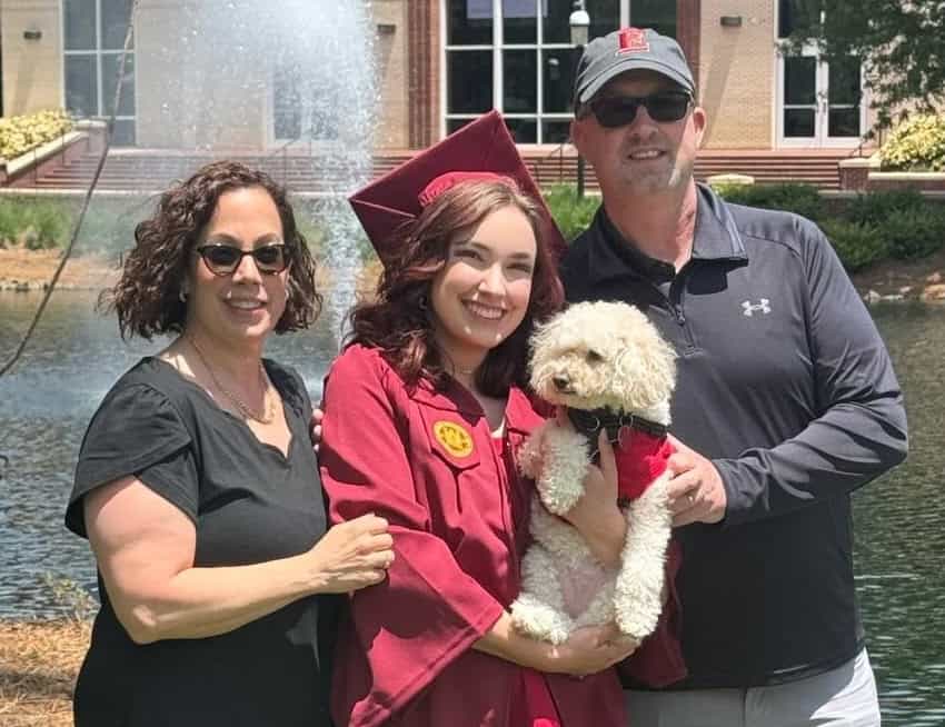 A woman in a maroon graduation gown and cap stands between her parents, holding a small, cream-colored dog in front of a fountain.