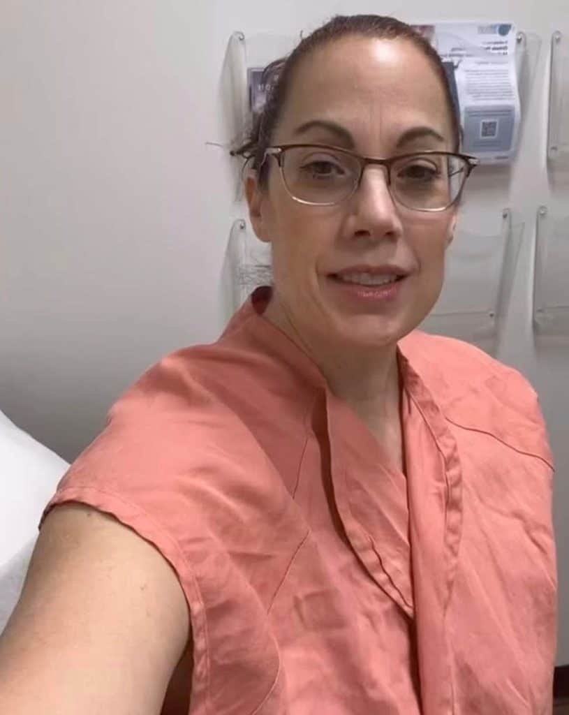 A woman with glasses wearing a peach-colored medical gown, reflecting on breast cancer survivor guilt.